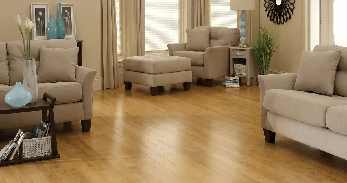 A living room with professionally installed bamboo flooring, beige sofas, a matching ottoman, a side table with a blue lamp, vases, framed pictures, and large windows with blinds and curtains in Austin, TX